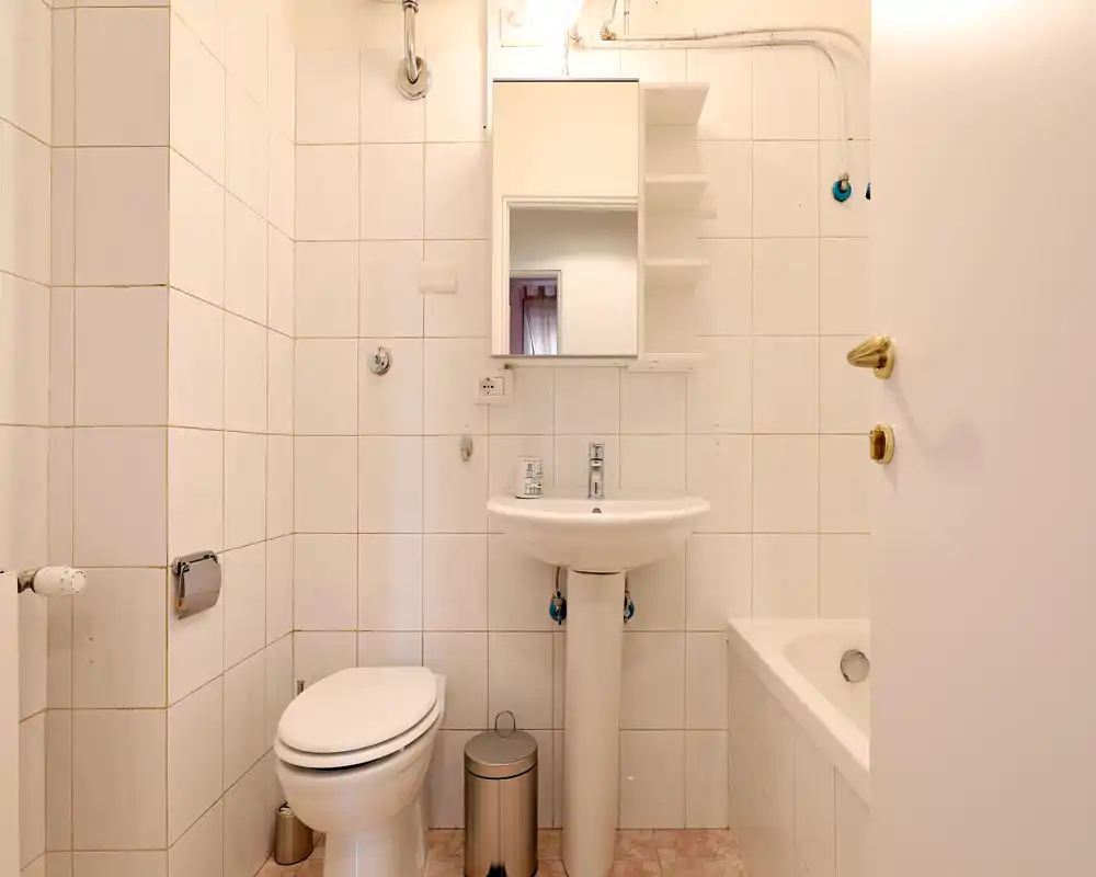 Bagno compatto e pulito con lavabo a colonna, WC, vasca e armadietto specchiato sopra il lavabo. Piastrelle bianche neutre e illuminazione soffusa donano un ambiente ordinato e funzionale.