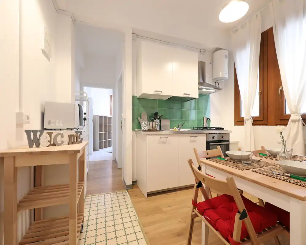 Cucina compatta e luminosa con paraschizzi piastrellato verde, mobili bianchi, piano cottura a gas e un piccolo tavolo da pranzo vicino alla finestra — accogliente e ben allestita per affitti brevi.