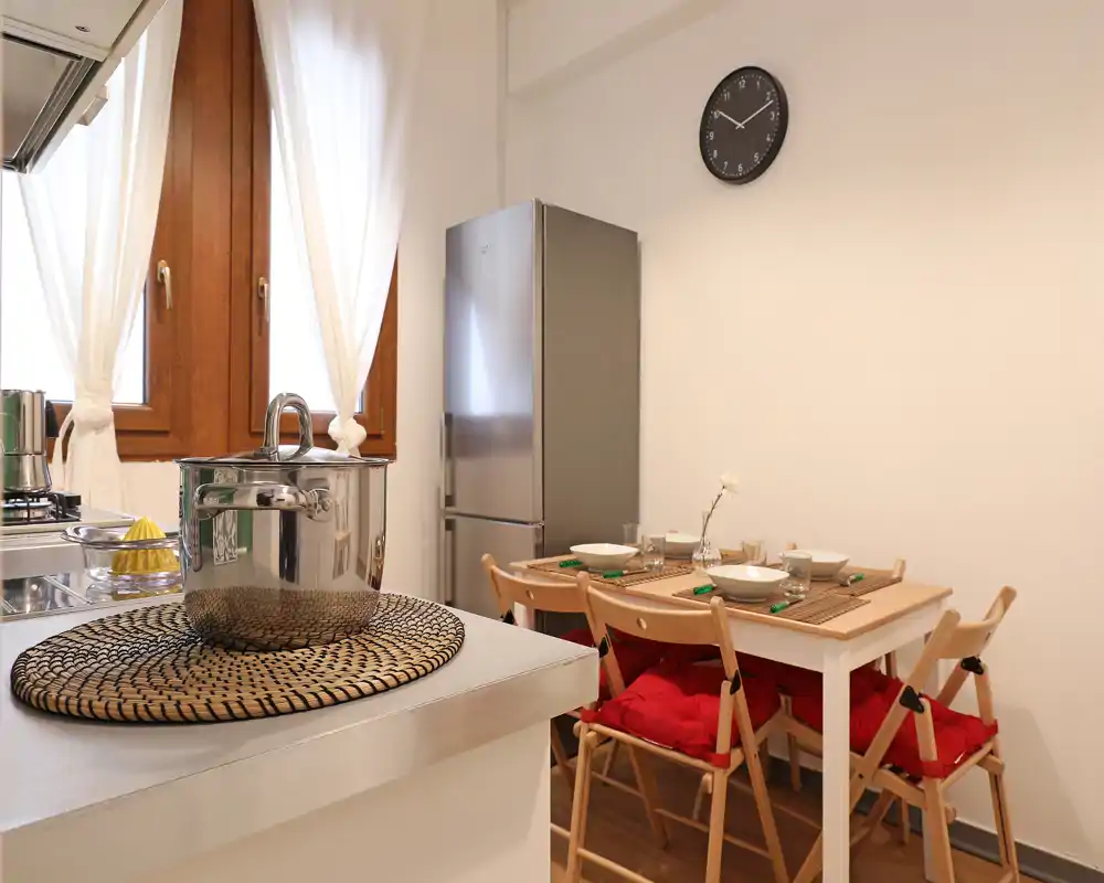 Cucina compatta e luminosa con un piccolo tavolo apparecchiato per quattro, frigorifero in acciaio e piano cottura visibile — uno spazio accogliente e ben illuminato ideale per pasti informali.