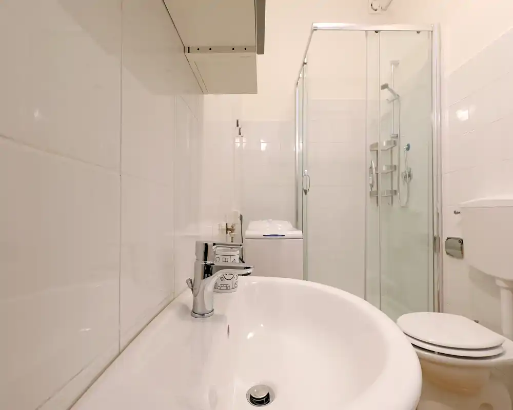 Bagno moderno e pulito con piastrelle bianche, lavabo in primo piano, box doccia in vetro e wc—luminoso e ben curato.
