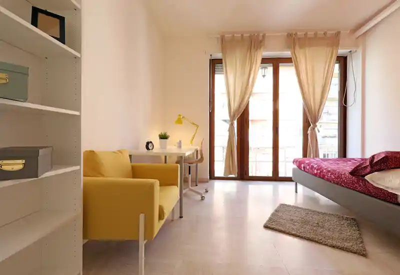 Bright, tidy bedroom with a single bed, a cozy armchair and a small desk facing a large balcony window that fills the room with natural light.