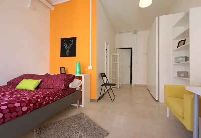 Bright, modern bedroom with a patterned double bed, colorful accent wall and cozy textiles. The room includes shelving, a small desk area and seating, offering a comfortable, well-lit living space ideal for short or long stays.