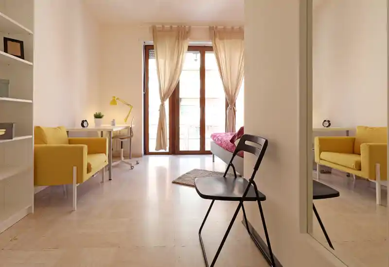 Bright, airy living area with two yellow armchairs, a small desk and a seating bed near a full-height window letting in generous natural light.