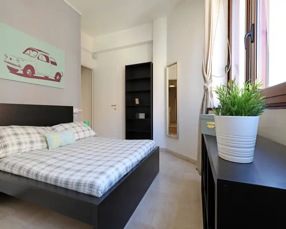Camera da letto moderna e luminosa con letto in legno scuro su pedana, biancheria a quadri e una grande finestra che illumina l'ambiente. Mensole minimal e uno specchio completano lo spazio arioso e ben arredato, perfetto per annunci.