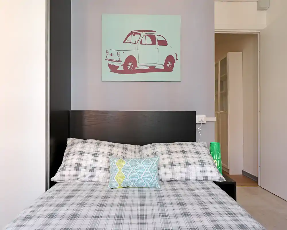 Camera da letto luminosa e ben composta con letto matrimoniale completo, biancheria a quadri e un cuscino decorativo sotto una stampa di un'auto sopra la testiera.