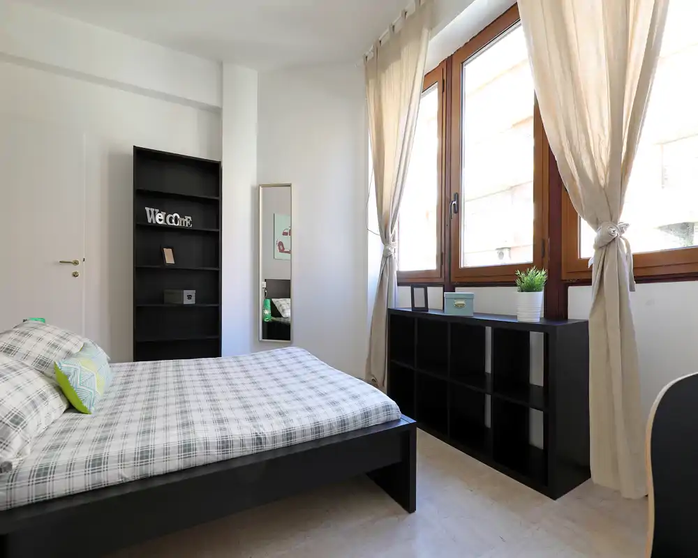 Camera da letto luminosa e moderna con letto matrimoniale ben rifatto, ampie finestre con tende beige e mobili neri con spazio contenitivo.