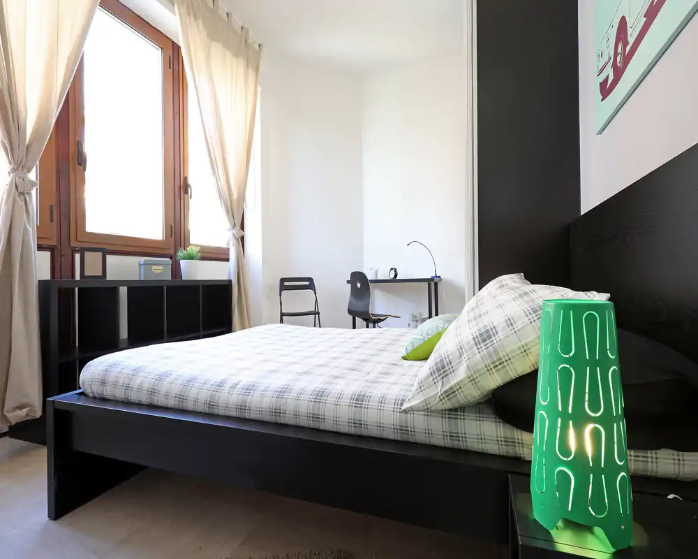 Camera da letto moderna e luminosa con letto basso matrimoniale, biancheria a quadri e una grande finestra che garantisce molta luce naturale. Una scrivania e mensole rendono la stanza funzionale per lavoro e riporre oggetti.