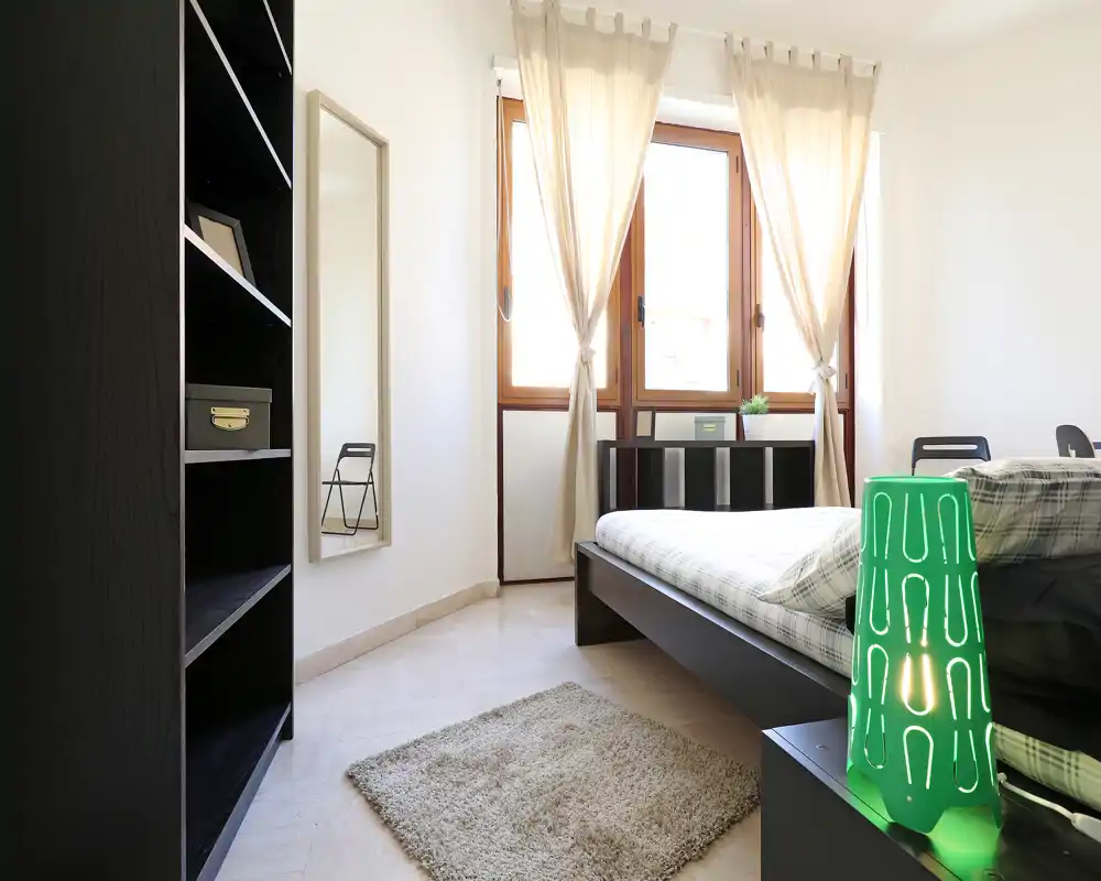 Camera da letto luminosa e moderna con letto basso, morbido tappeto e luce naturale dalle ampie finestre; decor minimal e lampada verde sul comodino rendono l'ambiente accogliente.