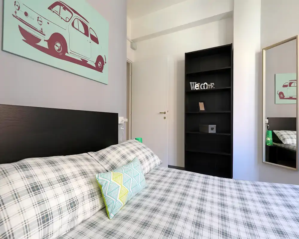 Camera da letto moderna e luminosa con ampio letto in primo piano, biancheria a quadri, cuscino decorativo e quadro alle pareti; scaffale pulito e specchio a figura intera.