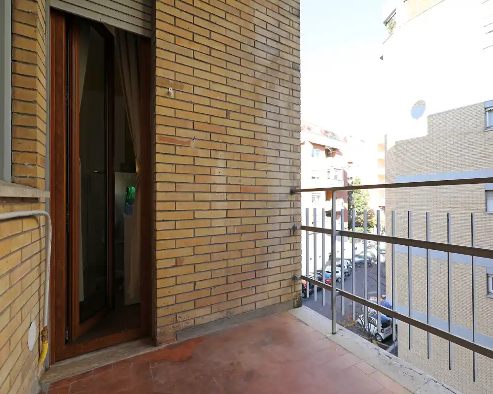 Piccolo balcone privato con pavimento piastrellato, parete in mattoni e ringhiera metallica con vista su una via tranquilla — luce naturale abbondante e accesso diretto dall'interno.