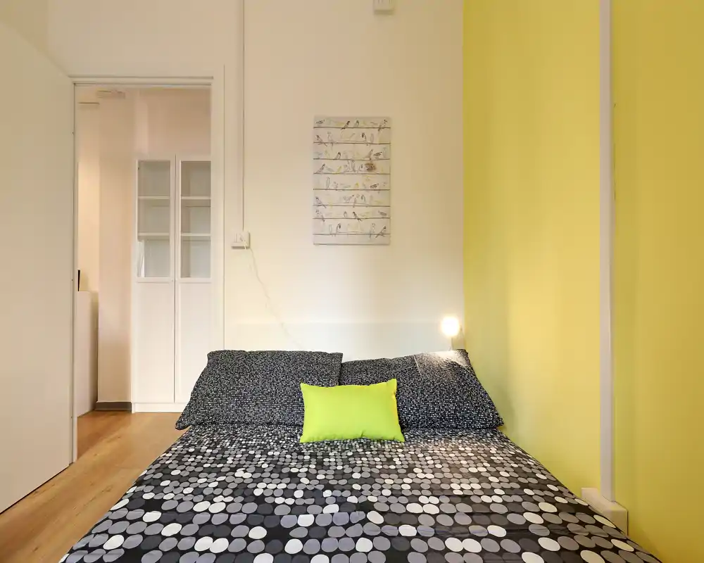 Camera da letto moderna e luminosa con matrimoniale a fantasia, cuscino verde lime e parete gialla vivace; ambiente ordinato e ben illuminato ideale per annunci.