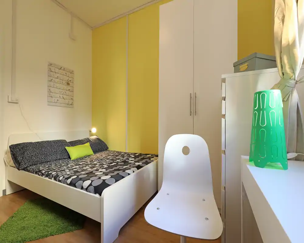 Camera da letto moderna e accogliente con letto bianco, biancheria a fantasia e cuscino verde. Armadio a muro, scrivania con lampada e luce naturale calda completano l'ambiente.