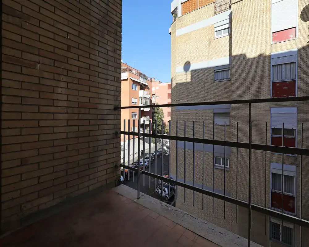 Piccolo balcone urbano che si affaccia su edifici residenziali e una strada; ringhiera in metallo, pareti in mattoni e pavimento in cotto, cielo sereno ben visibile.