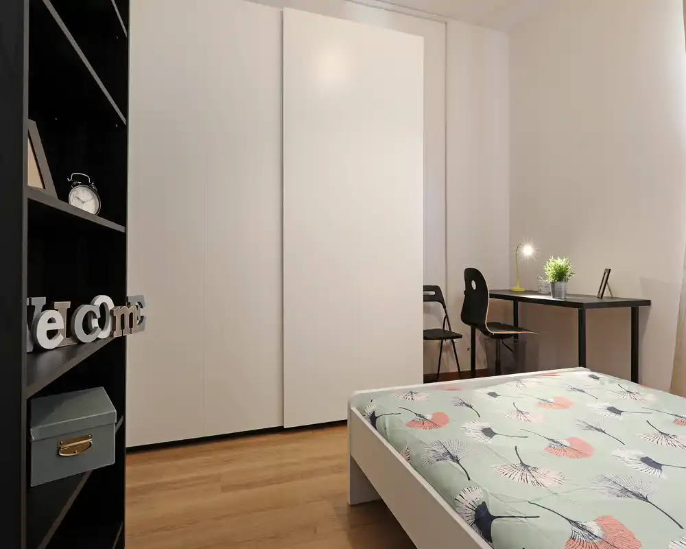 Camera da letto moderna e minimal con letto matrimoniale dal copriletto stampato, ampia armadiatura bianca e una scrivania compatta con sedia e lampada — luminosa, ordinata e ideale per affitti a breve termine.