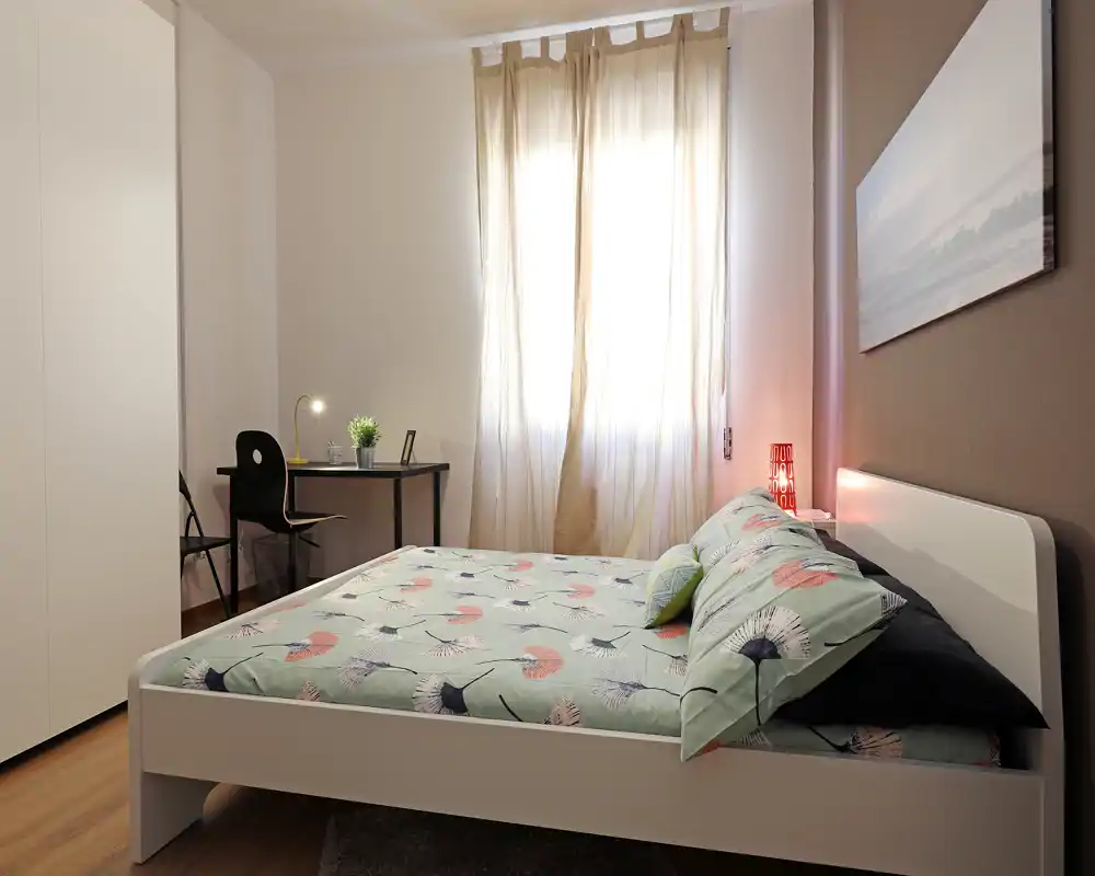 Camera da letto luminosa e ben arredata con un letto completo in primo piano, luce naturale morbida dalla finestra con tenda, una lampada da comodino e una piccola scrivania — accogliente e pronta per gli ospiti.