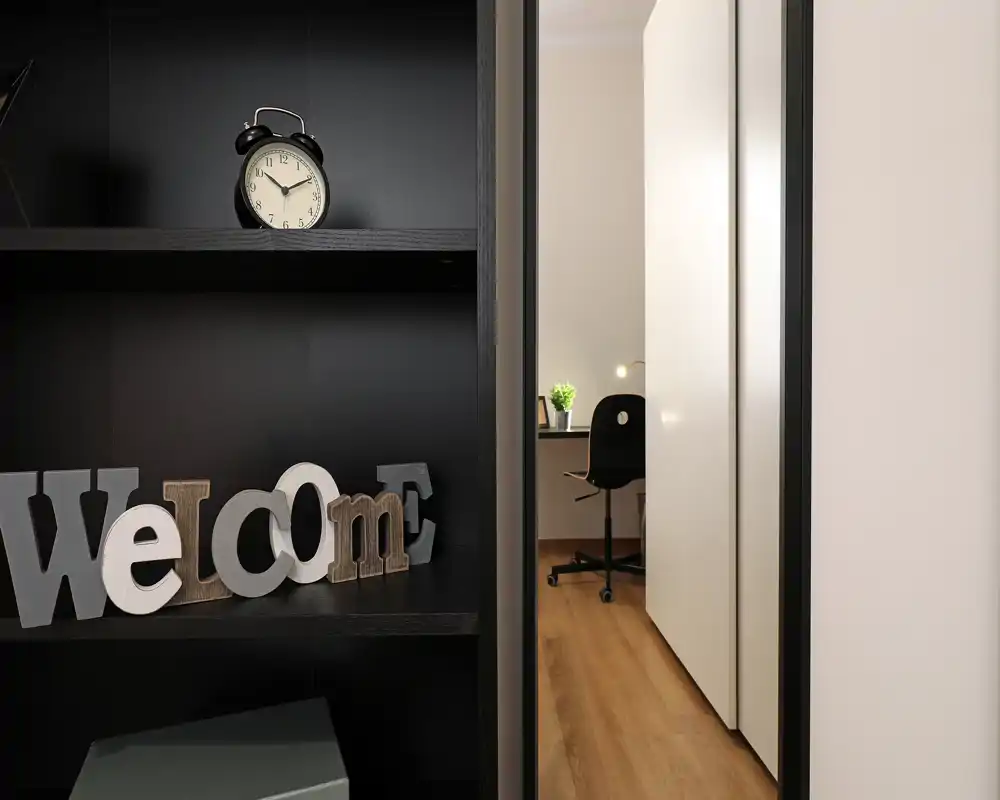 Ingresso di un soggiorno moderno e ordinato con mensole decorative e scorcio di una piccola postazione di lavoro. Il pavimento in legno caldo e la decorazione "Welcome" creano un'atmosfera accogliente.