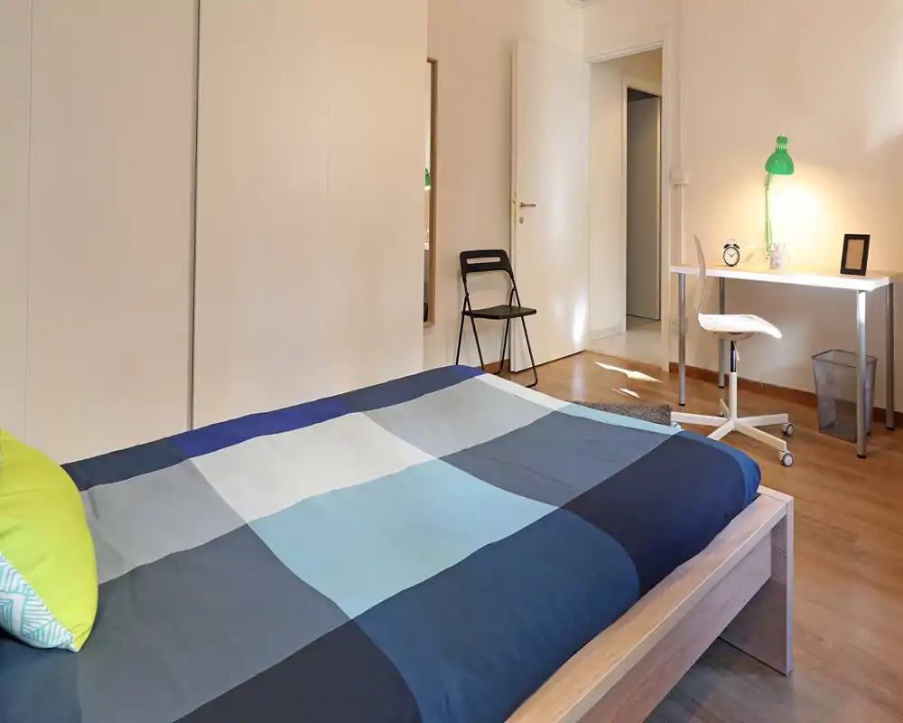 Camera da letto moderna e luminosa con un ampio letto in legno in primo piano, armadio minimale e una scrivania ordinata con lampada verde — ideale per studenti o professionisti.