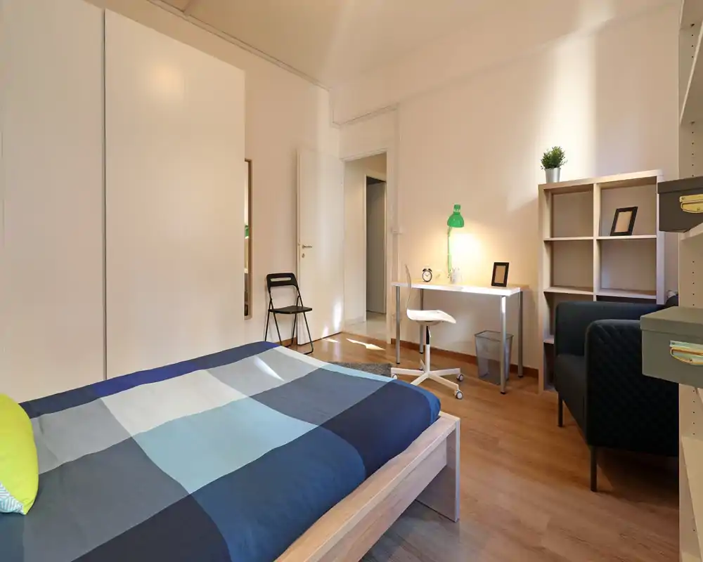 Camera da letto luminosa e ben arredata con letto in legno moderno, tessili confortevoli e un angolo studio con scrivania e lampada — ideale per una persona o una coppia.