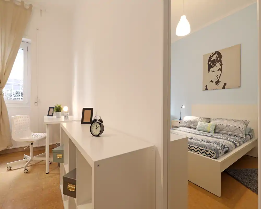 Dormitorio luminoso y estiloso con cama doble, ropa de cama estampada, lámpara de mesita y cuadro en la pared; se ve también una zona de trabajo ordenada contigua.