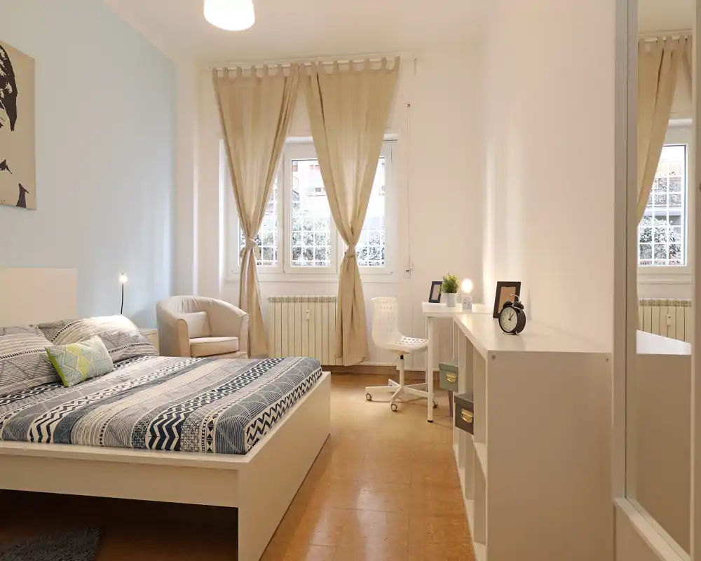 Dormitorio luminoso y bien decorado con una gran cama blanca con ropa de cama estampada, un sillón acogedor junto a la ventana y una pequeña zona de escritorio — ideal para estancias cómodas.