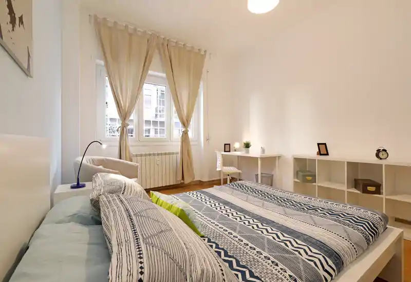 Dormitorio luminoso y bien decorado con cama doble grande, ropa de cama con estampado, escritorio y estanterías en la pared — ideal para estancias cómodas.