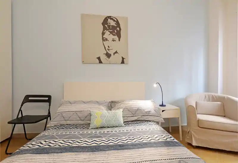 Dormitorio luminoso y bien decorado con cama grande, ropa de cama estampada, mesita con lámpara y un sillón acogedor — ideal para estancias tranquilas.