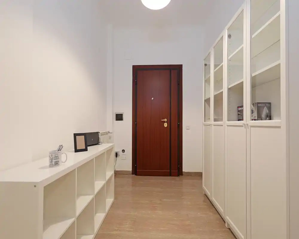 Vestíbulo ordenado y bien iluminado con puerta de entrada de madera, armarios blancos y estantería abierta — estilo minimalista y limpio, ideal como entrada de alquiler.