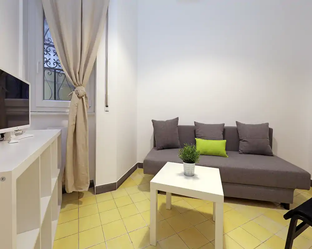 Zona de estar compacta y moderna con sofá gris, cojines de acento y una pequeña mesa de centro blanca. Paredes neutras y luz natural suave crean un espacio cuidado y apto para alquileres temporales.