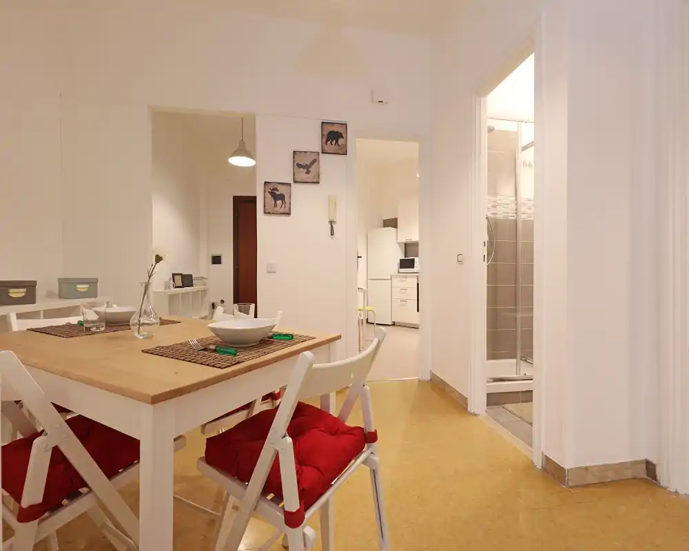 Zona de comedor bien decorada con mesa de madera, sillas plegables con cojines rojos y preparaciones para comer; espacio acogedor y ordenado que comunica con la cocina y el baño.