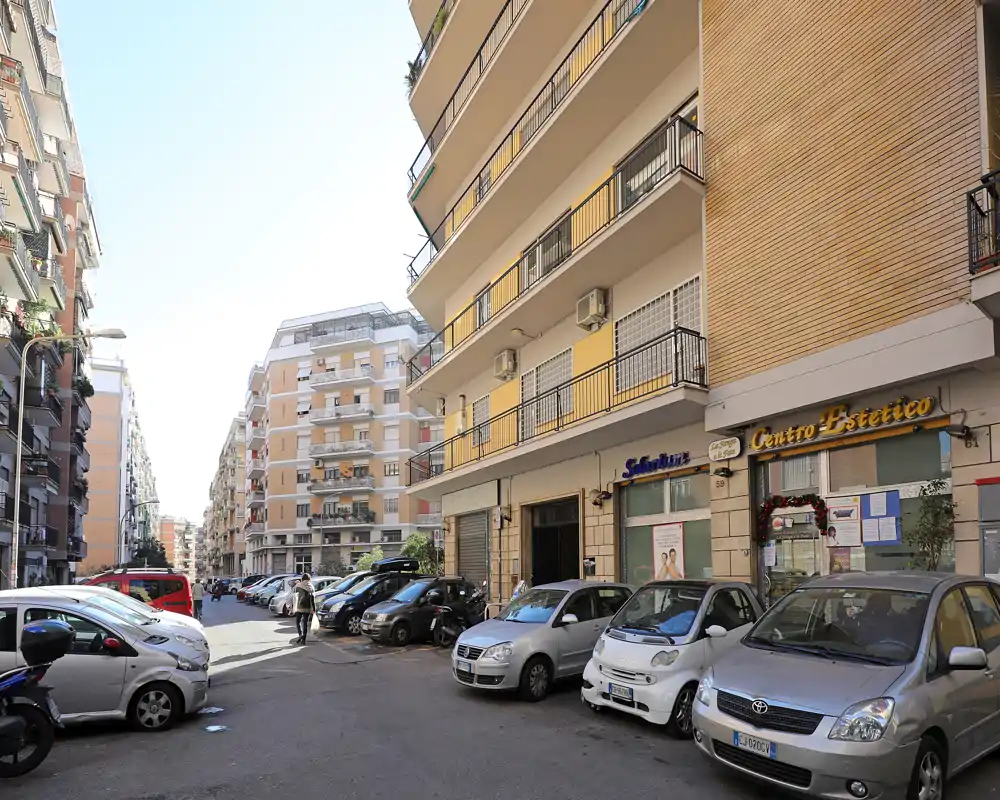 Vista de una calle urbana soleada con coches aparcados y edificios residenciales de altura media; locales en planta baja y balcones que transmiten ambiente de barrio.