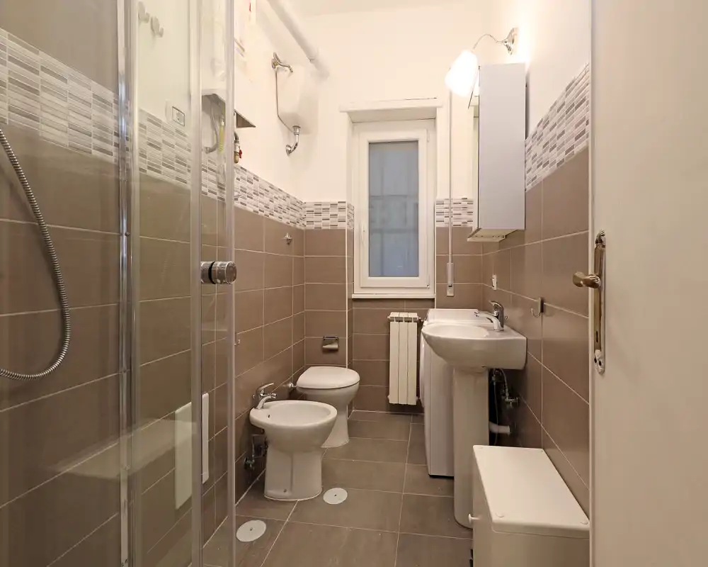 Baño moderno y bien iluminado con ducha, bidé, inodoro y lavabo, paredes alicatadas y una pequeña ventana que aporta luz natural.