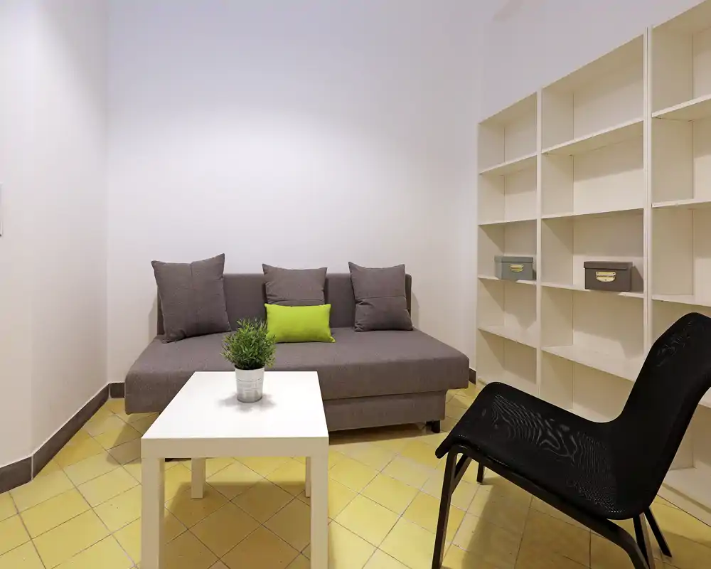 Espacio de estar compacto y moderno con sofá gris, cojín verde, mesa blanca y estanterías — luminoso, ordenado y apto para un piso pequeño.