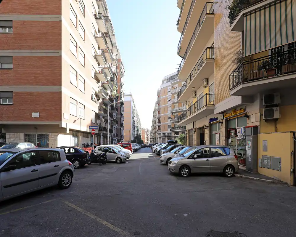 Vista de una calle residencial soleada con coches aparcados y edificios de altura media a ambos lados; luz diurna clara y un pequeño comercio en planta baja visible.