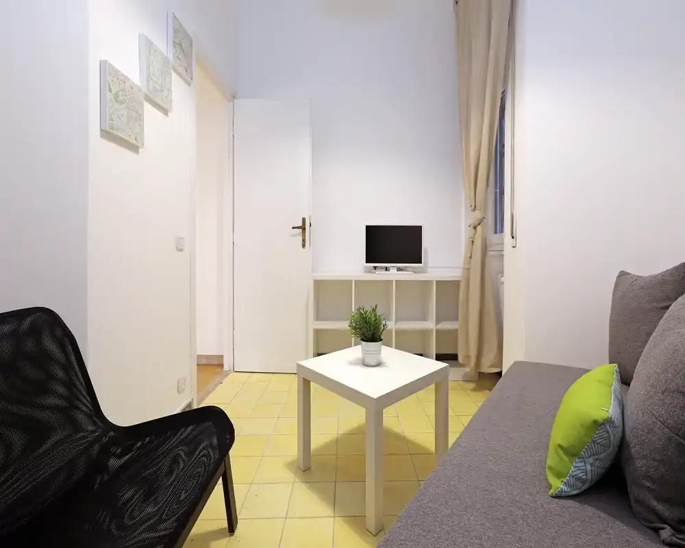 Salón pequeño y luminoso con sofá gris, cojines, mesa de centro blanca y una televisión compacta sobre estantería. Estilo minimalista y limpio, suelo de baldosas y luz natural desde la ventana.