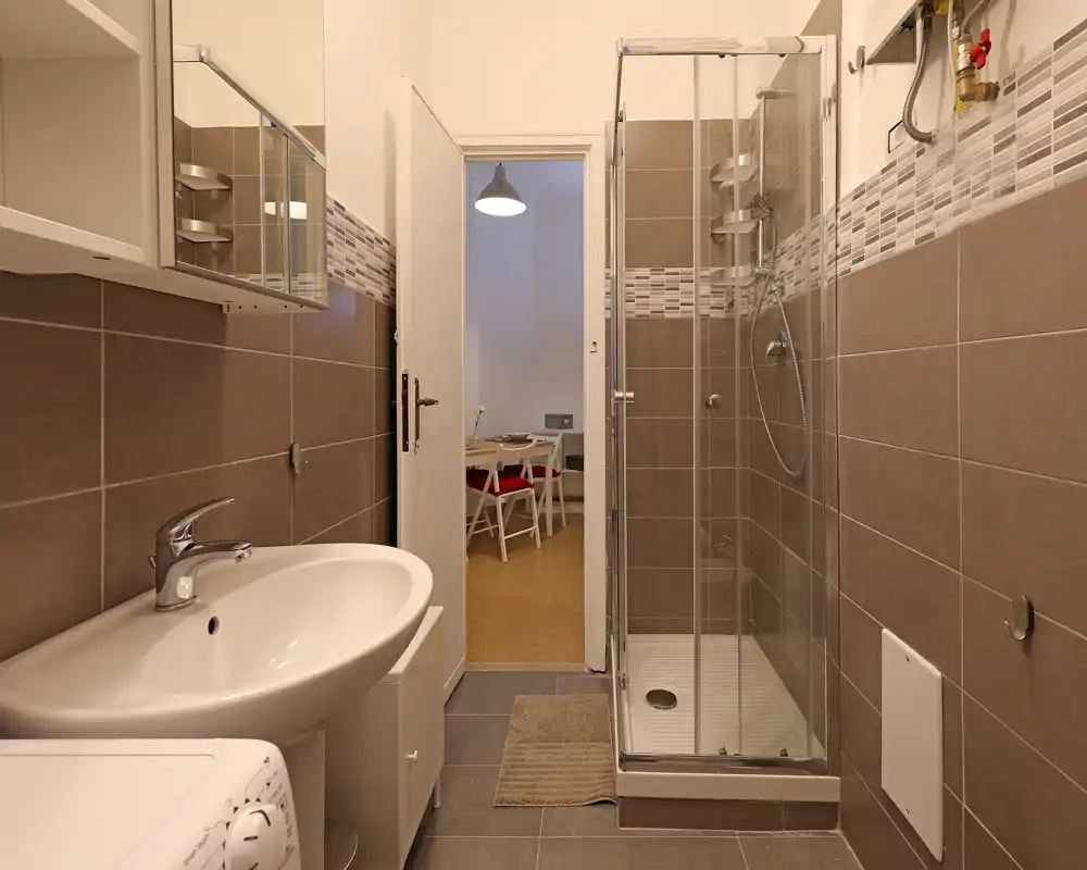 Baño moderno y luminoso con plato de ducha de vidrio en esquina, azulejos y lavabo blanco; la puerta abierta muestra una zona de comedor/trabajo.
