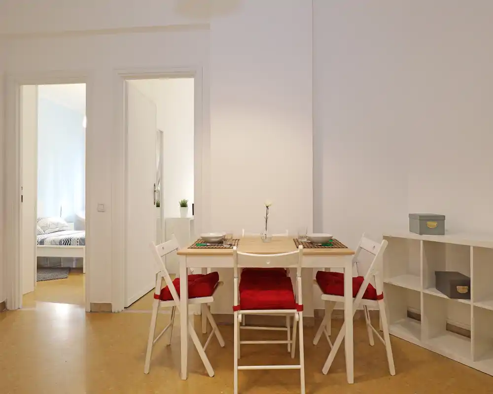 Zona de comedor bien iluminada con mesa de madera sencilla, cuatro sillas plegables blancas con cojines rojos, manteles individuales y un pequeño jarrón — ordenada, minimalista y acogedora para comidas informales.