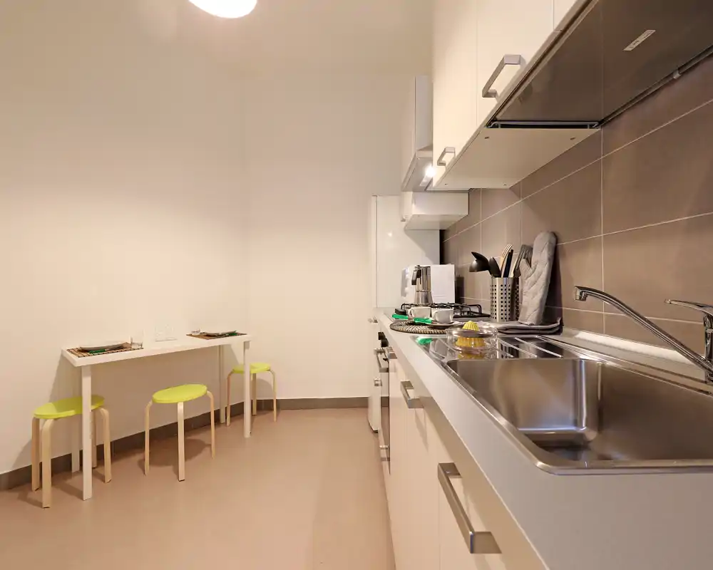 Cocina moderna y compacta con una larga encimera, fregadero de acero inoxidable, armarios integrados y una pequeña mesa con tres taburetes: práctica y ordenada para el uso diario.