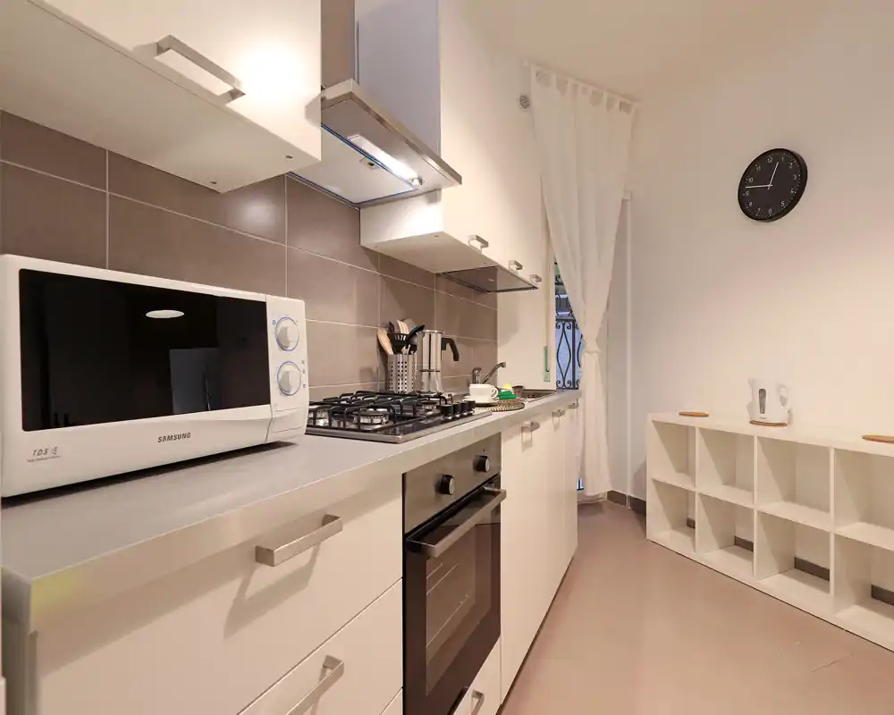 Cocina compacta y moderna con placa de gas, horno empotrado y microondas, encimeras luminosas y estantería ordenada — ideal para cocinar con eficiencia.