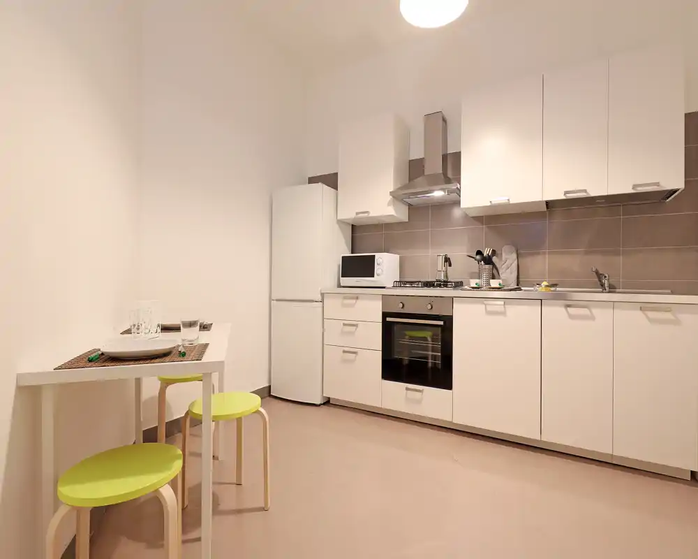 Cocina moderna y compacta con armarios blancos, horno empotrado, placa de gas, frigorífico y microondas. Una pequeña barra con taburetes verdes aporta un área para comer informal.