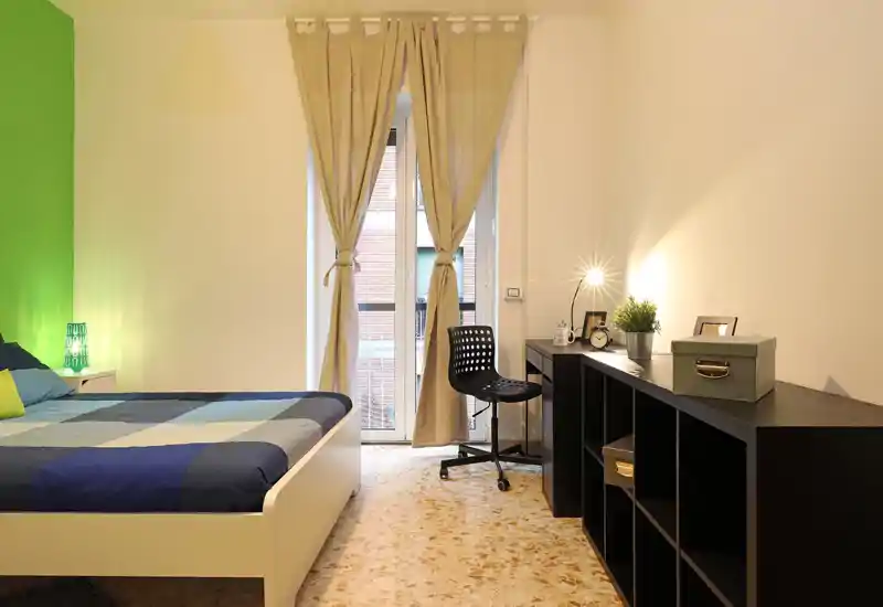 Dormitorio moderno y luminoso con una cama doble grande y ropa de cama a cuadros; un rincón de estudio con escritorio, silla y estanterías ocupa la pared derecha, y una ventana con cortinas claras aporta luz natural.