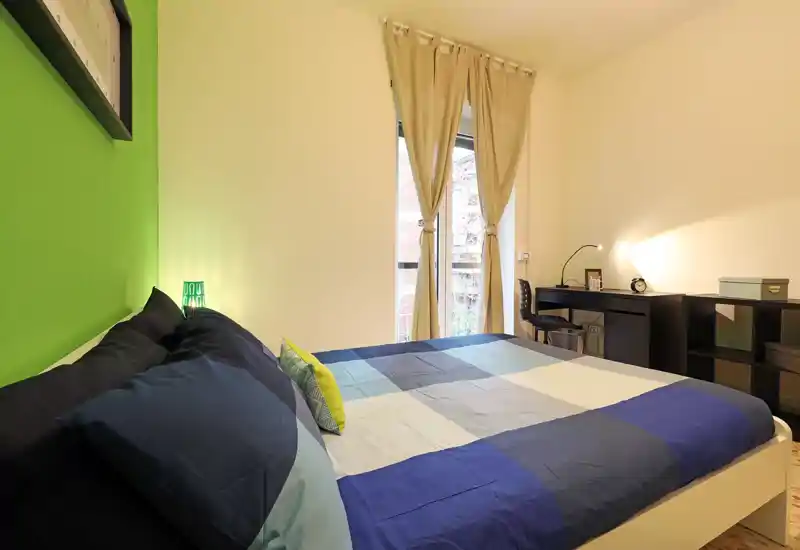 Dormitorio moderno y luminoso con cama doble grande con edredón azul y gris, pared verde de acento y escritorio junto a la ventana — ideal para descansar o teletrabajar.