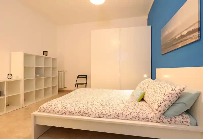 Dormitorio luminoso y ordenado con cama doble vestida con ropa de cama estampada, armario blanco y estanterías; pared azul de acento y decoración mínima para un aspecto moderno y fresco.