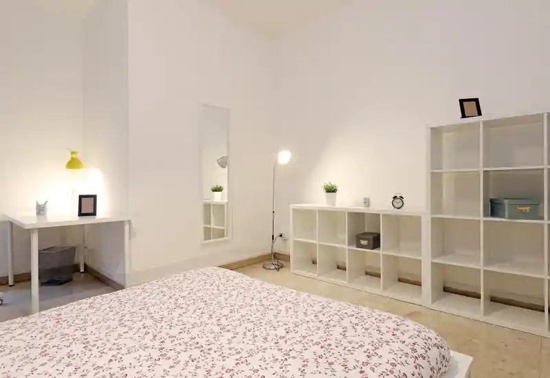 Dormitorio luminoso y minimalista con cama estampada en primer plano, estanterías blancas, un pequeño escritorio con lámpara amarilla y plantas decorativas — espacio limpio y acogedor, ideal para un anuncio.
