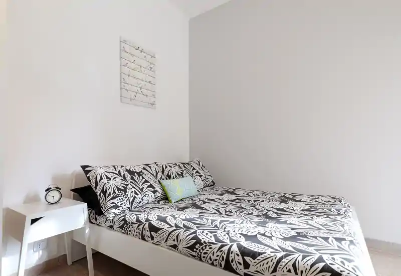 Dormitorio luminoso y minimalista con cama doble con ropa de cama de motivo botánico, mesita con despertador; paredes neutras y luz natural que crean un ambiente moderno y tranquilo.
