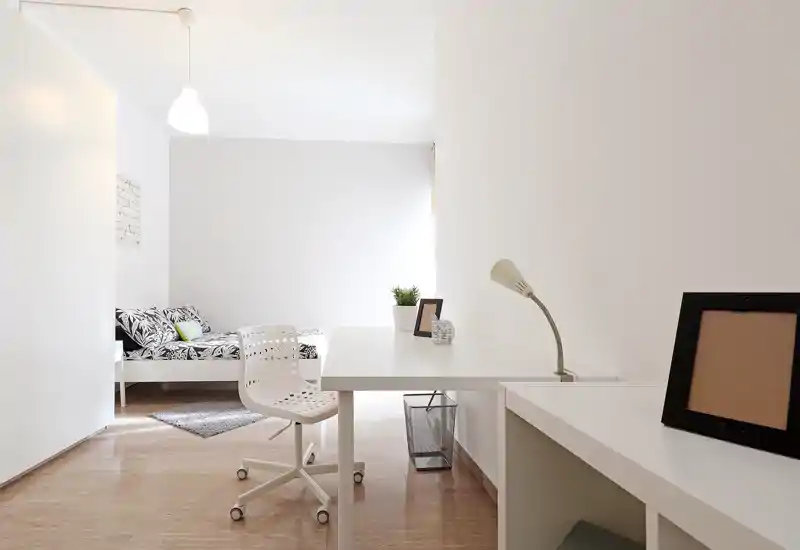 Dormitorio minimalista y bien iluminado con un escritorio blanco en primer plano y una cama individual estampada en el fondo. Estilo moderno y limpio con una alfombra pequeña y cojines decorativos.