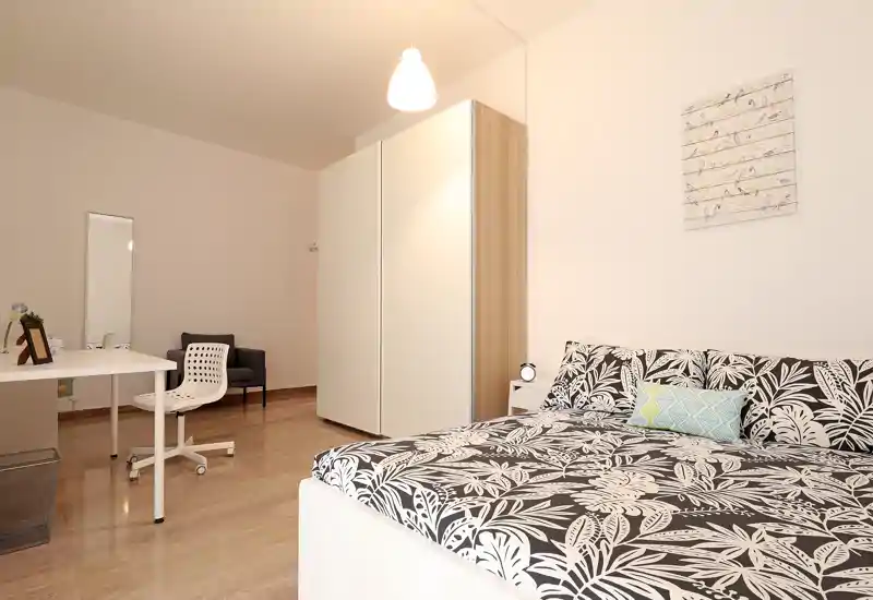 Dormitorio moderno y luminoso con cama doble con estampado, mesita y armario. La habitación está bien puesta en escena con escritorio y silla, suelo de madera y una lámpara colgante suave.