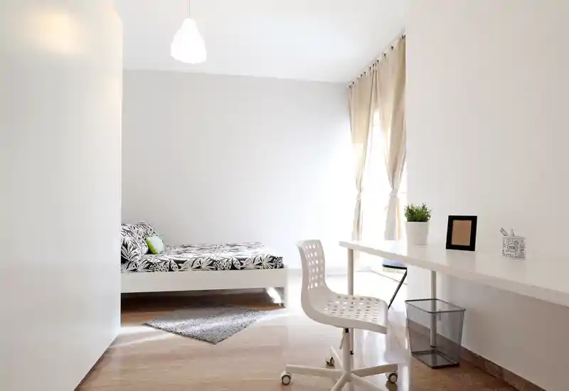 Dormitorio luminoso y minimalista con cama baja blanca, ropa de cama con estampado, zona de escritorio y luz natural de una ventana alta. Líneas limpias y tonos neutros que crean un ambiente sereno y aireado para anuncios de alquiler.