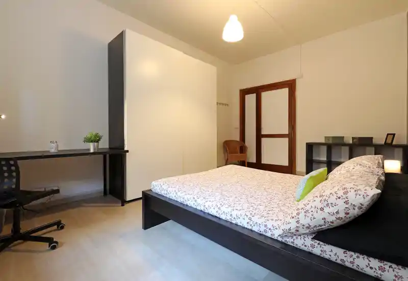 Dormitorio moderno y luminoso con cama doble sobre estructura oscura, armario minimalista, escritorio y estanterías — cómodo para estancias cortas o largas.