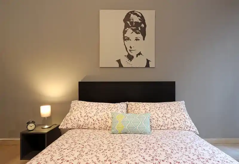 Dormitorio luminoso y ordenado con ropa de cama estampada, cojines decorativos y una lámpara de mesita que crea un ambiente cálido y acogedor.