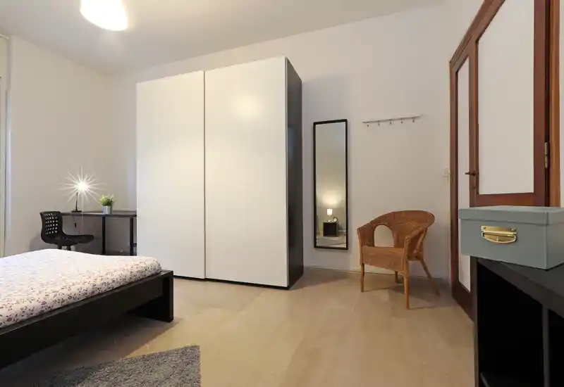 Dormitorio moderno y luminoso con cama visible, armario blanco, escritorio y silla de mimbre; tonos neutros y líneas limpias para un ambiente acogedor.
